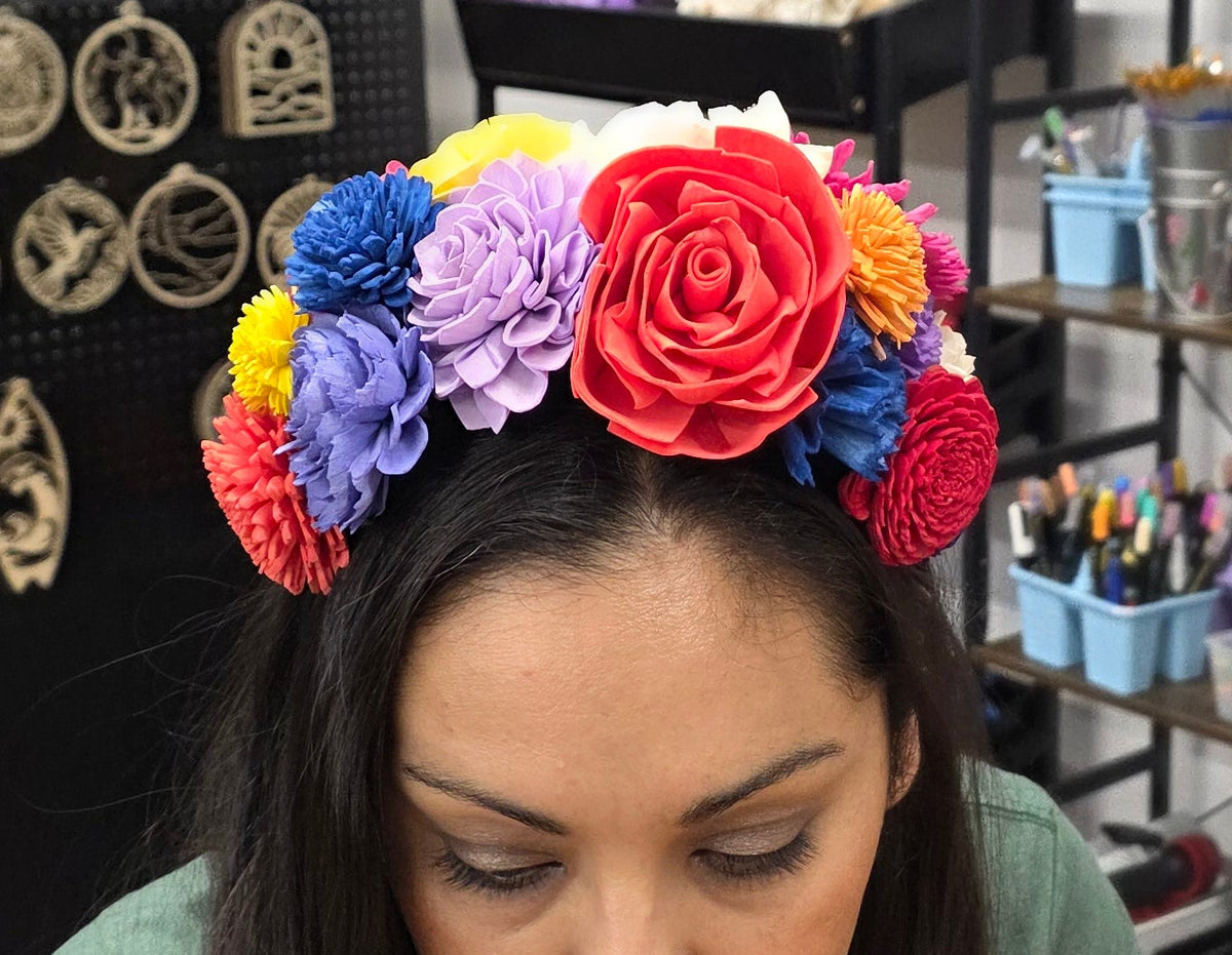 A woman with long dark hair wearing a colorful artificial flower crown sits at a table in a craft room. The background shows shelves with supplies and two floral wreaths hanging on the wall.