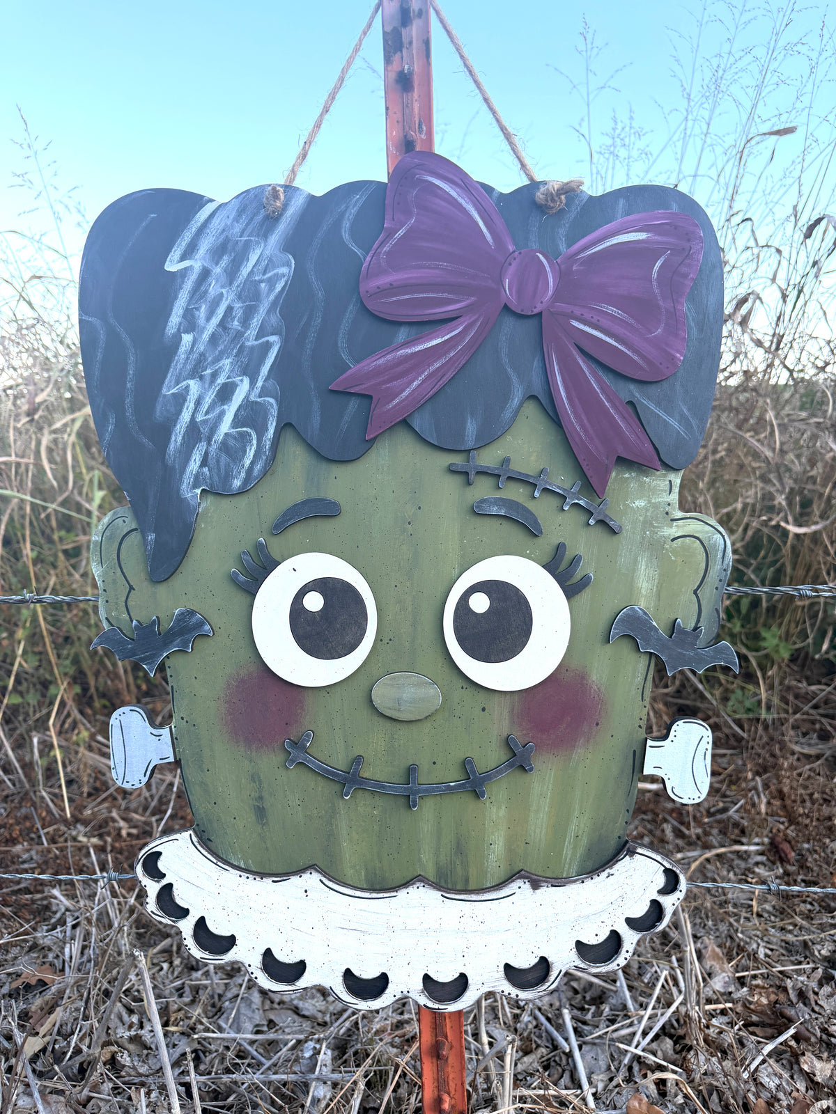A decorative hanging sign shaped like a Frankenstein-style monster head, with dark green face paint, a purple bow, black hair, large white eyes, and a white ruffled collar, photographed outdoors in a field.