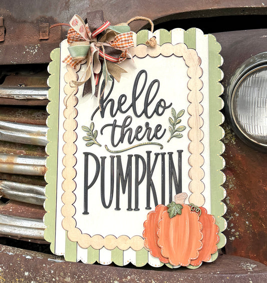 A decorative autumn sign reading 'hello there PUMPKIN' with a scalloped cream border, green stripes, a small orange pumpkin, and a ribbon bow, placed against the front of a rusted vintage truck outdoors.
