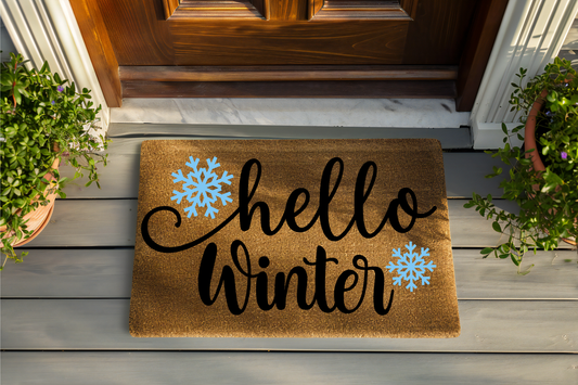 Doormat with 'hello winter' text and snowflake designs on a wooden floor.