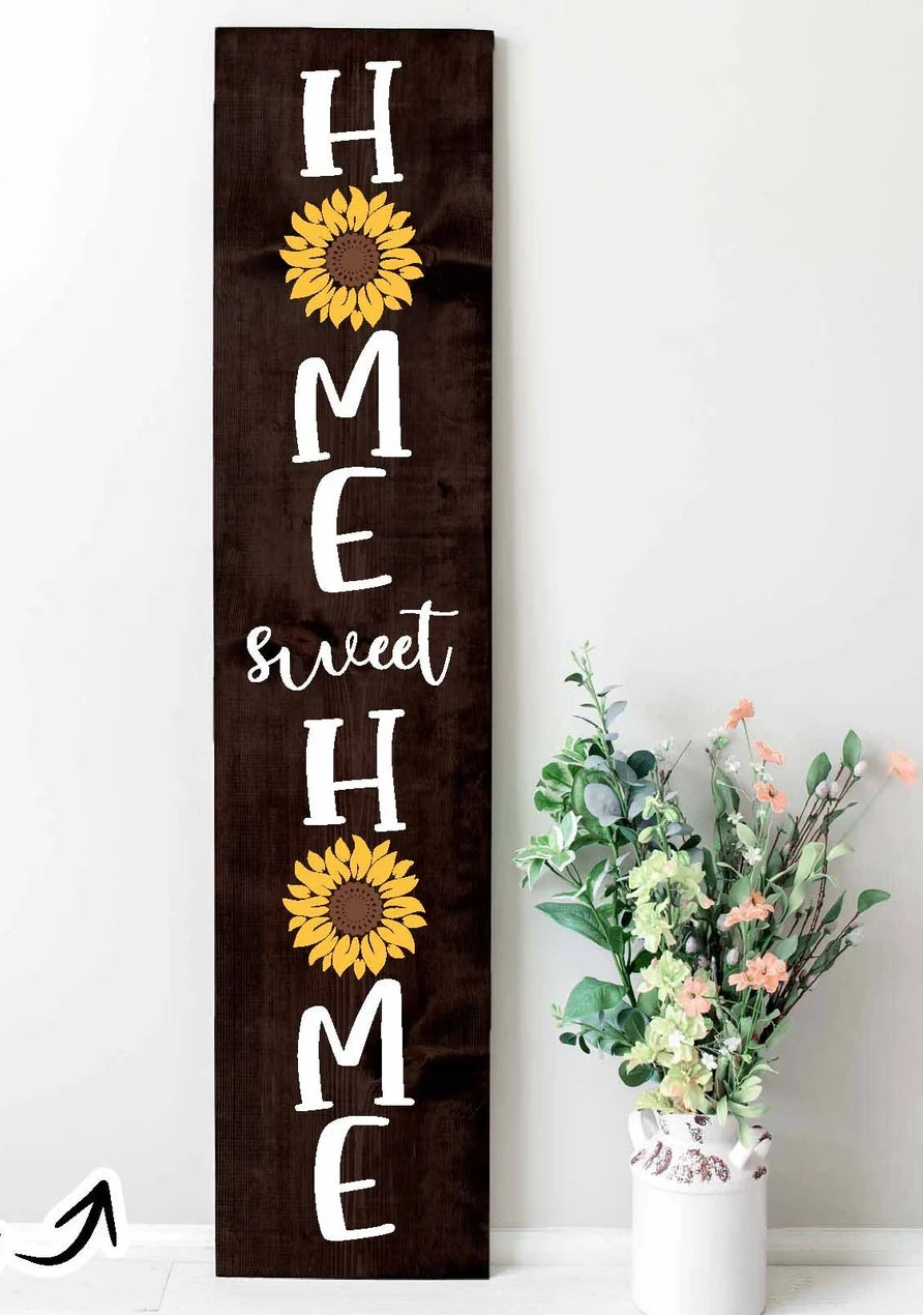 A tall rectangular dark brown wooden sign with the phrase 'HOME sweet HOME' in white lettering and two yellow sunflower graphics, displayed against a white wall next to a white vase with flowers. A small 'Project idea' text overlay appears in the bottom-left corner.