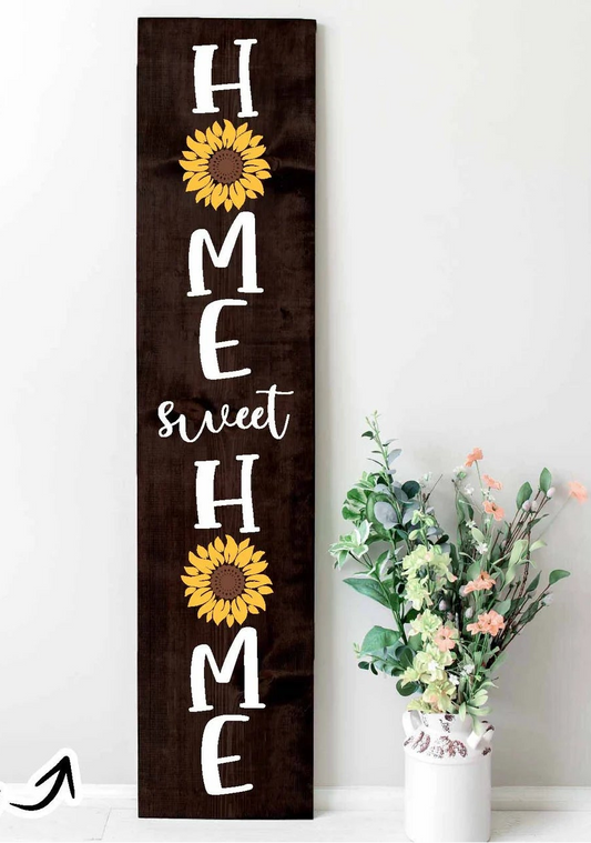 A tall rectangular dark brown wooden sign with the phrase 'HOME sweet HOME' in white lettering and two yellow sunflower graphics, displayed against a white wall next to a white vase with flowers. A small 'Project idea' text overlay appears in the bottom-left corner.