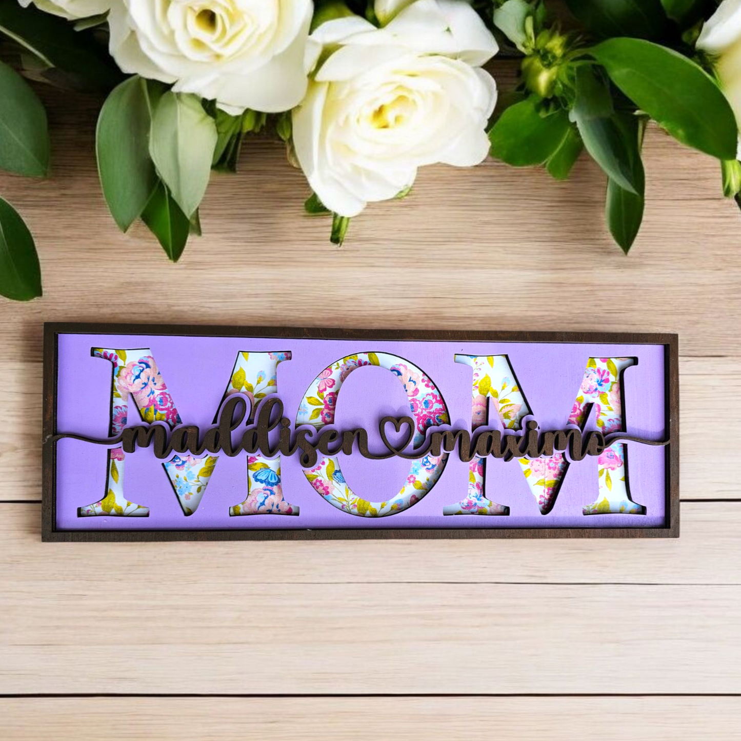 A personalized shelf sitter sign that spells out 'MOM' with each letter styled with a different floral pattern, mounted on a purple background, placed on a wooden surface with white roses and green leaves in the background.
