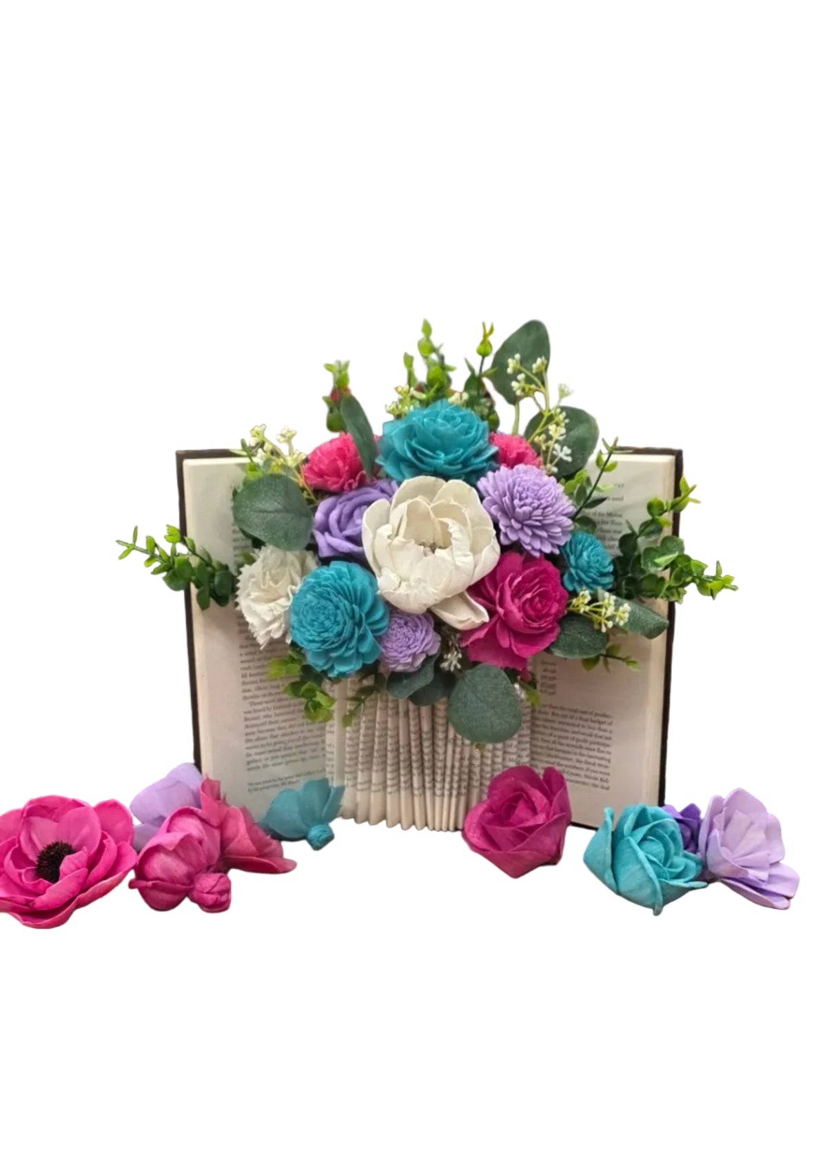 A colorful bouquet of artificial paper flowers in shades of pink, blue, purple, and white, arranged on a wooden table inside a workshop or studio.