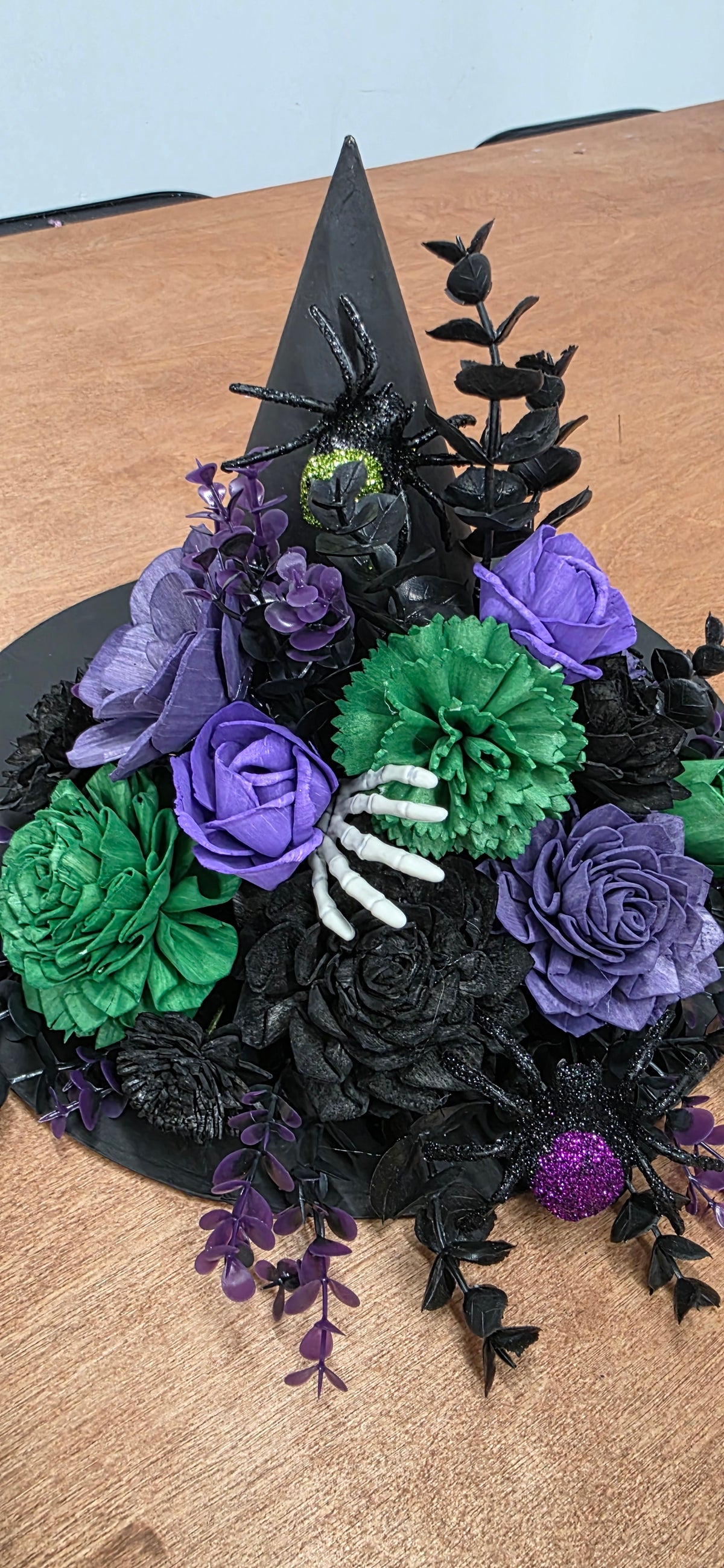 A black witch hat centerpiece decorated with purple and green fabric flowers, a white spider hand, and black spiderwebs, displayed on a wooden table.