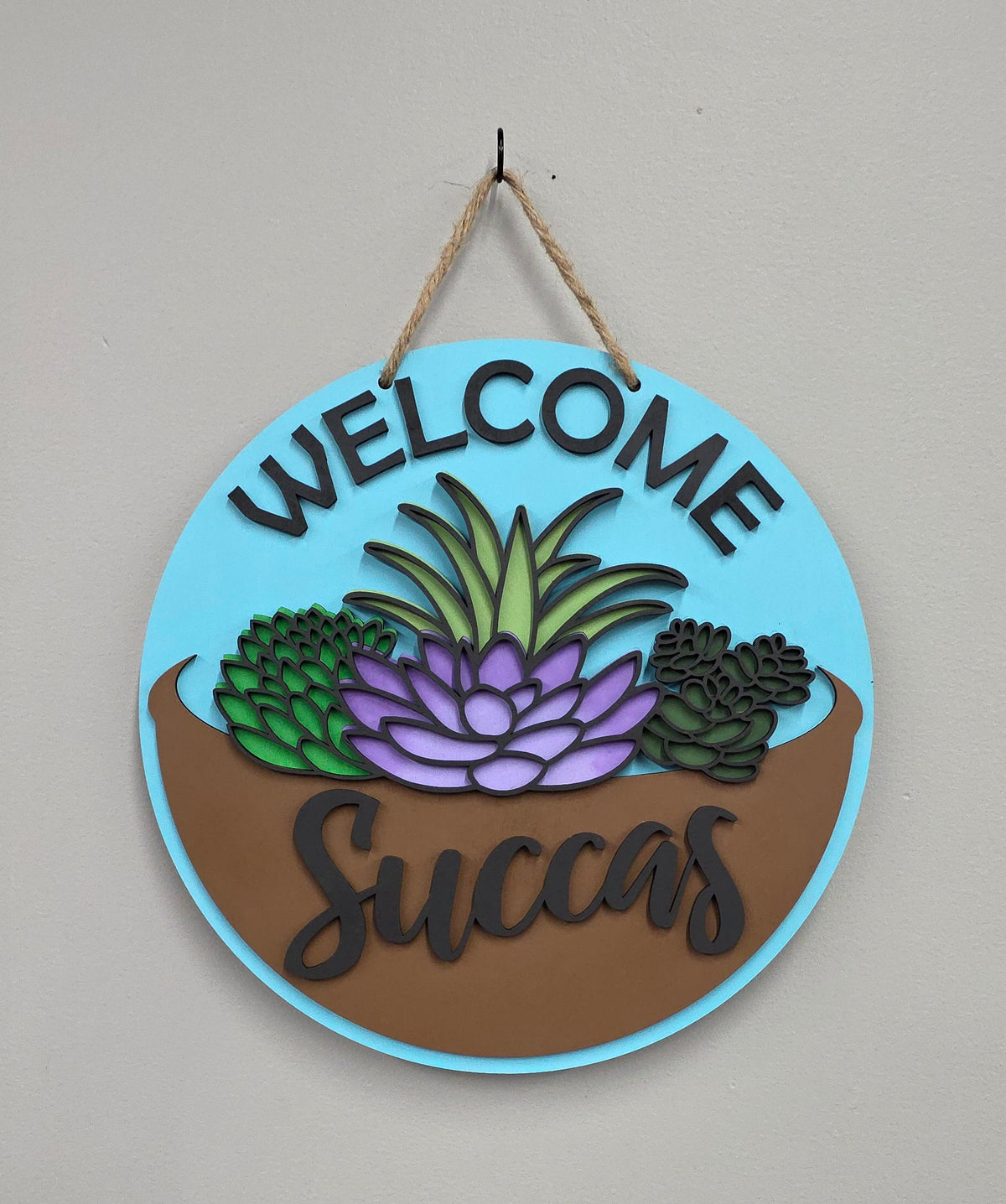 A round hanging wall sign with a light blue border, brown lower section, and colorful succulent illustration. The sign reads “WELCOME Succas” in raised lettering and hangs from a rope against a plain light gray wall.